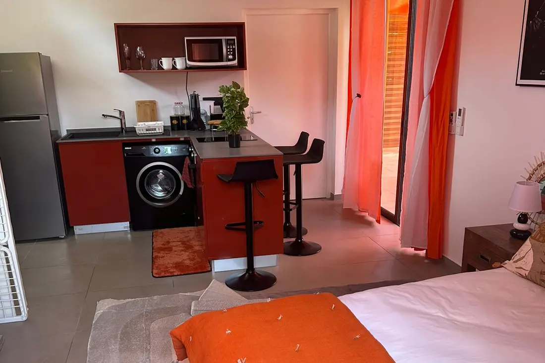 Studio de la Résidence Francis : kitchenette rouge avec lave-linge et micro-ondes, bar, tabourets noirs et lit au premier plan