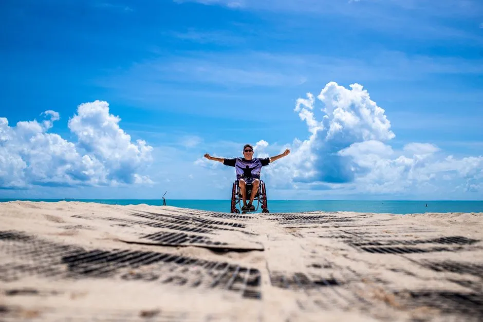 Personne en fauteuil roulant bras leves sur un tapis d'acces menant a une plage tropicale au bord d'une mer turquoise