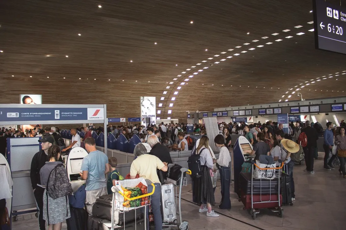 Hall d'enregistrement d'un aéroport français vu depuis le sol : files de passagers avec chariots à bagages devant les comptoirs, bornes libre-service en premier plan, panneaux « Carte d'embarquement » et « Étiquette bagage », plafond en bois clair et écrans d'information suspendus