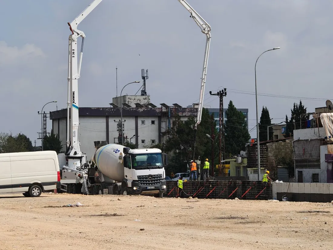 Chantier de gros œuvre photographié au niveau du sol : une toupie à béton et une pompe à béton à flèche déployée stationnées sur une plateforme de terre, trois ouvriers en gilet orange et casque jaune travaillant le long d'un coffrage, bâtiments et lignes électriques en arrière-plan