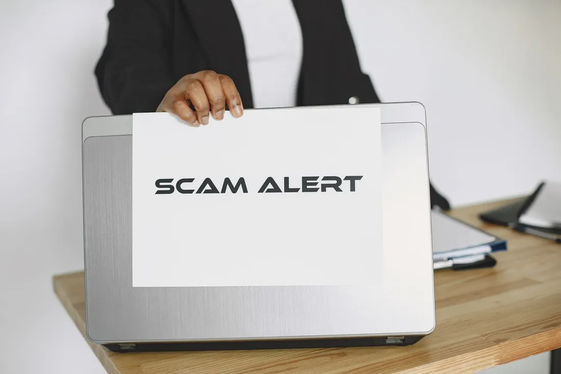 Personne en tenue de bureau brandissant une feuille marquée « SCAM ALERT » (alerte à l'arnaque) au-dessus d'un ordinateur portable, sur un bureau