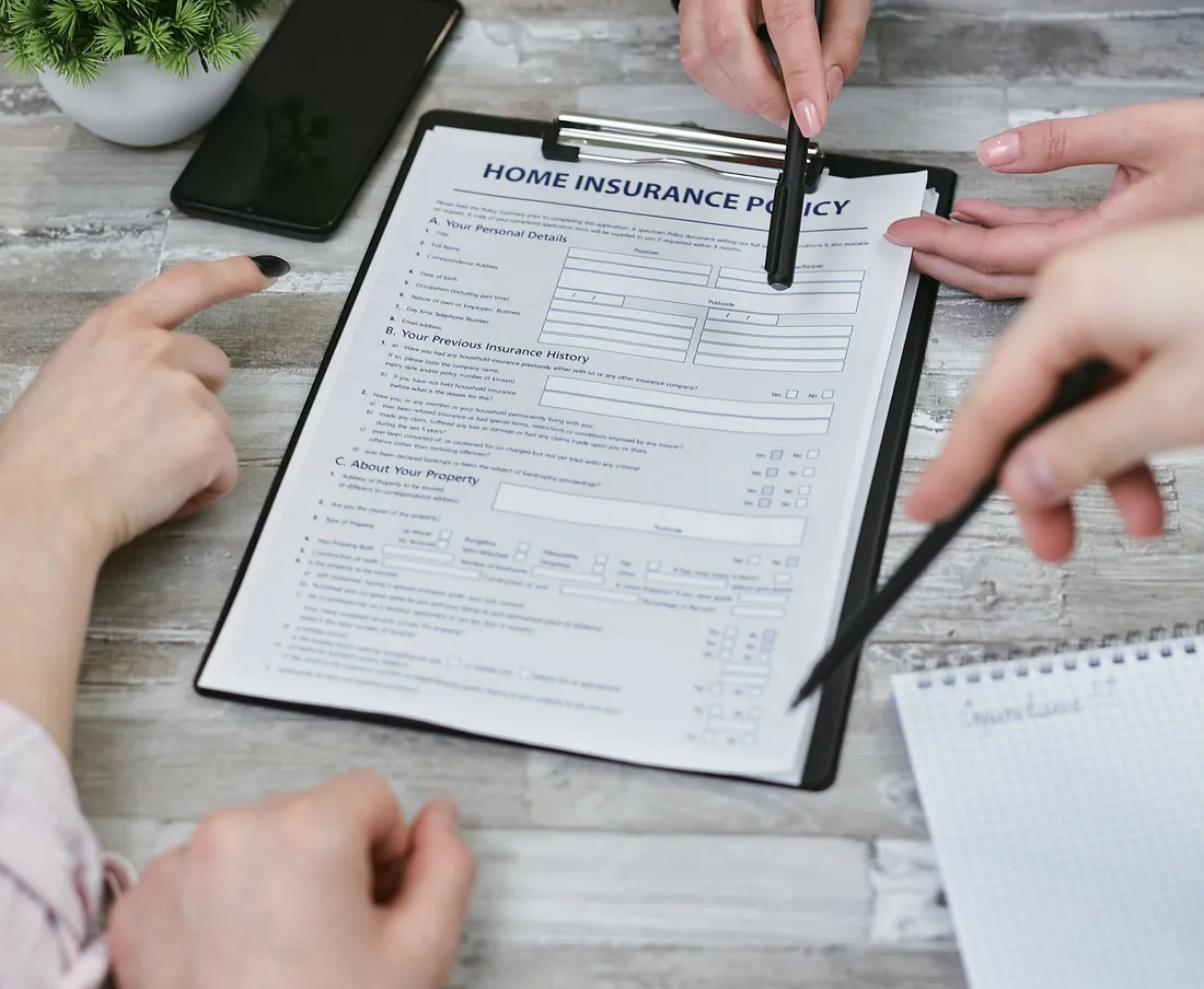 Trois personnes penchées autour d'un formulaire de contrat d'assurance habitation posé sur une planche à pince, stylos à la main, l'une pointant du doigt une rubrique du document