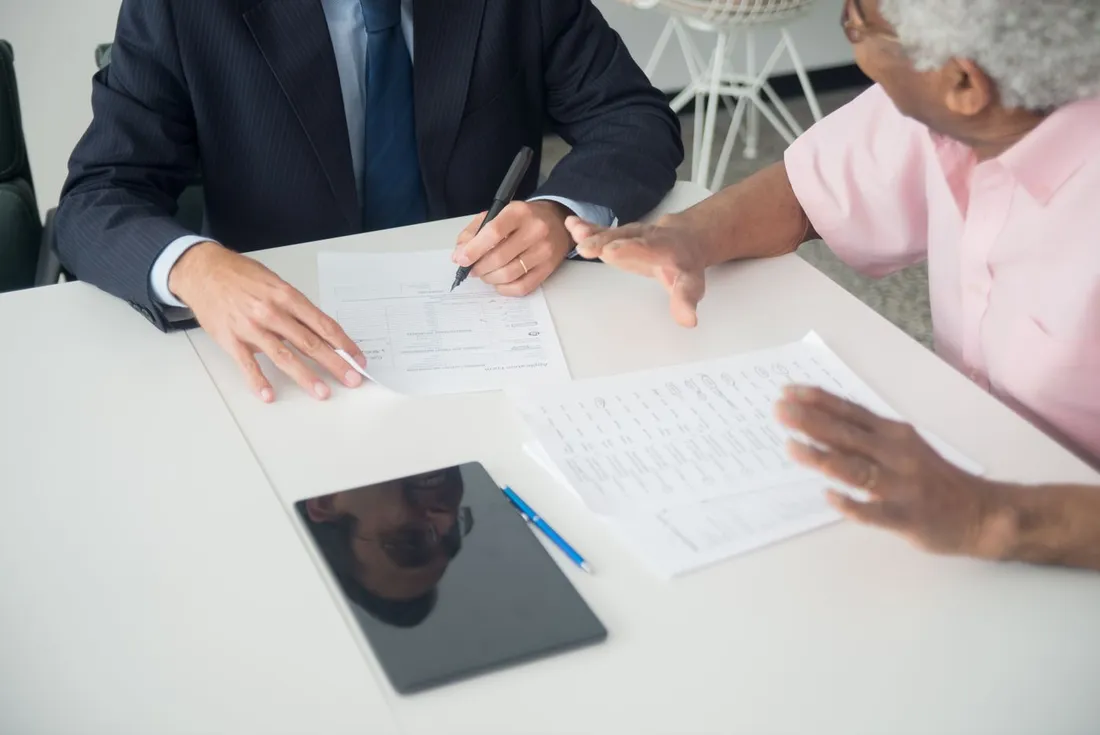 Un conseiller en costume remplit un formulaire de contrat sur un bureau, face à un client âgé qui tient une seconde page du dossier