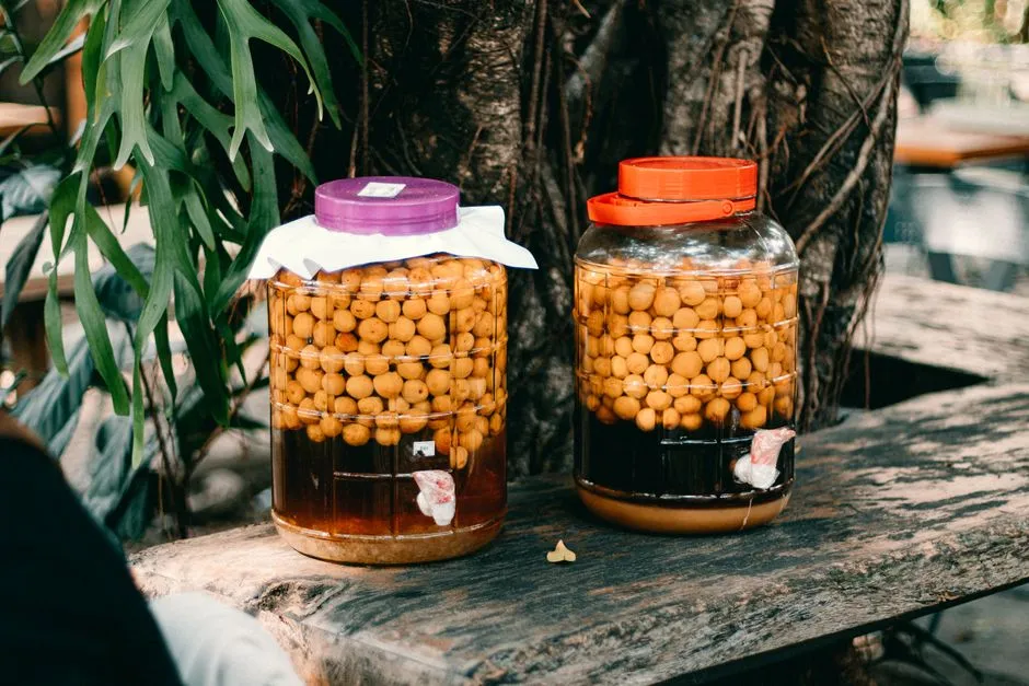 Deux grandes bonbonnes en verre remplies de fruits en cours de macération dans du rhum, posées sur une table en bois dans un cadre tropical