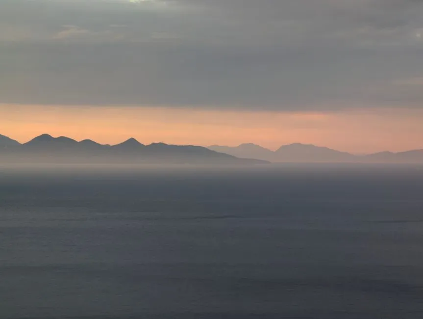 Littoral aux reliefs montagneux estompés par une brume épaisse couleur sable, ciel laiteux et horizon voilé, illustrant un épisode de brume de sable du Sahara au-dessus de la mer