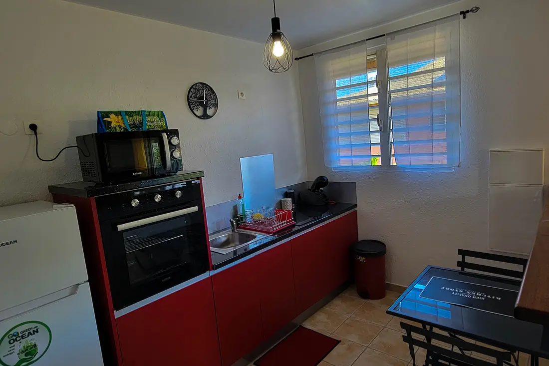 Cuisine rouge d’un logement avec four noir, micro-ondes, horloge murale et bar en bois donnant sur la fenêtre