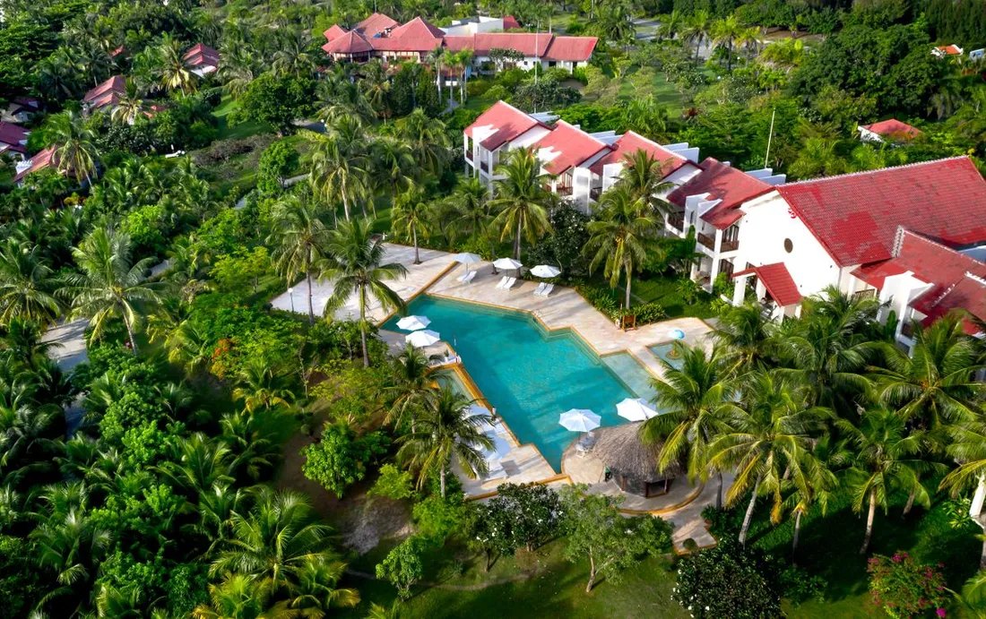 Vue aerienne d'une residence touristique tropicale avec piscine entouree de palmiers et de bungalows, illustrant un bien immobilier en location saisoniere