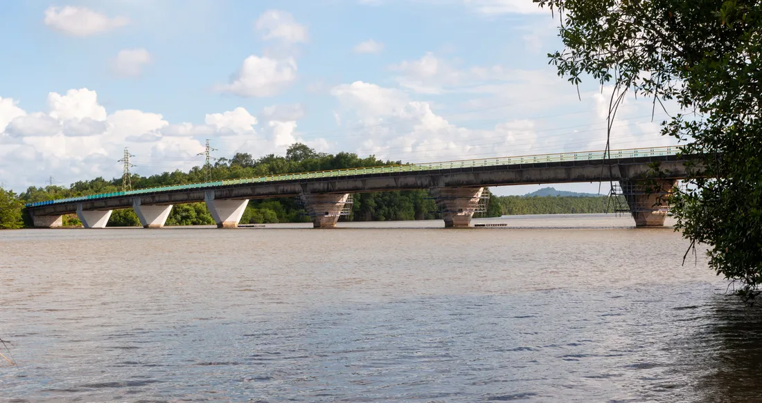 Pont routier en béton franchissant le fleuve Kourou, vu depuis la rive : le tablier bordé d'une glissière verte, la mangrove et la forêt sur l'autre berge, ciel de traîne