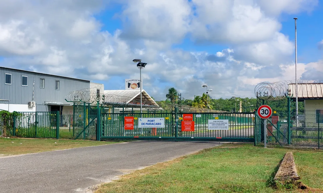 Entrée du port de Pariacabo à Kourou : grille métallique fermée, clôture surmontée de barbelés, panneau « Port de Pariacabo » et panneau de zone protégée interdisant l'accès sans autorisation