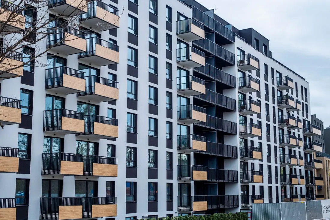 Façade d'un immeuble résidentiel neuf photographiée depuis la rue : sept étages d'appartements aux balcons habillés de bois clair et garde-corps métalliques noirs, larges baies vitrées, et une palissade de chantier encore en place au pied du bâtiment à droite
