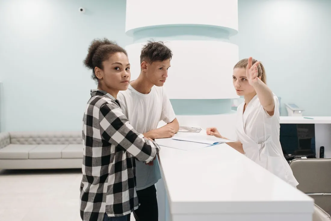 Deux patients accoudés au comptoir d'un accueil hospitalier ; une soignante en blouse blanche, de l'autre côté du comptoir, leur indique une direction du bras tendu