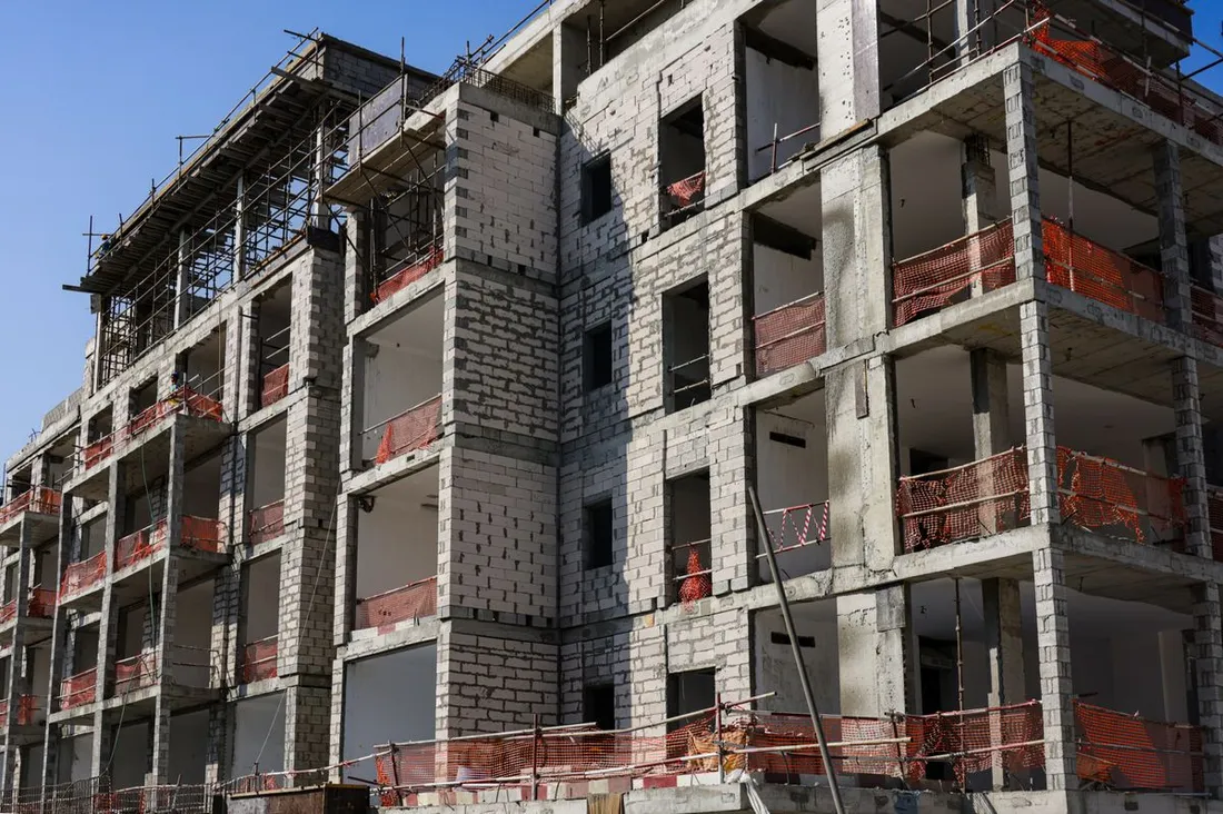 Bâtiment de plusieurs étages en cours de construction photographié depuis la rue : structure poteaux-poutres en béton et murs en blocs, ouvertures encore béantes, filets orange de protection aux balcons, échafaudages sur le dernier niveau, ciel bleu