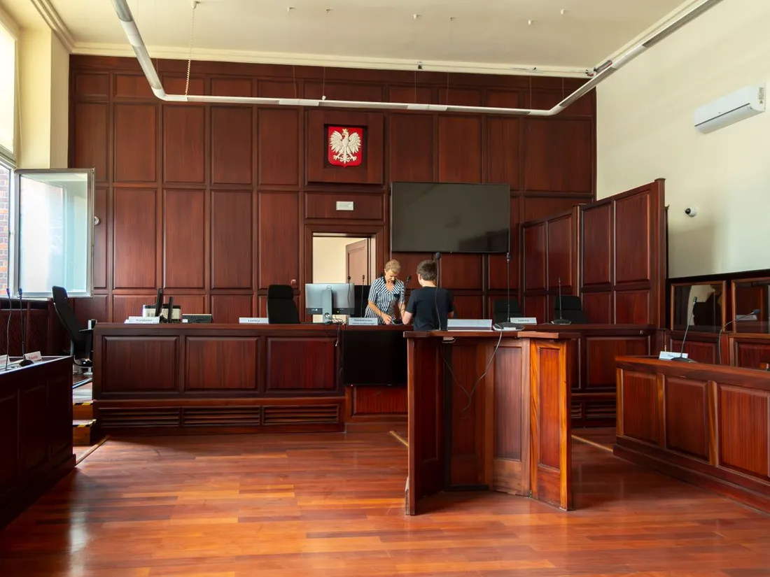 Salle d'audience lambrissée de bois sombre photographiée depuis le fond de la salle : l'estrade et les sièges des magistrats au fond, une barre en bois au centre, des pupitres équipés de micros de part et d'autre, un écran mural, deux personnes debout près de l'estrade