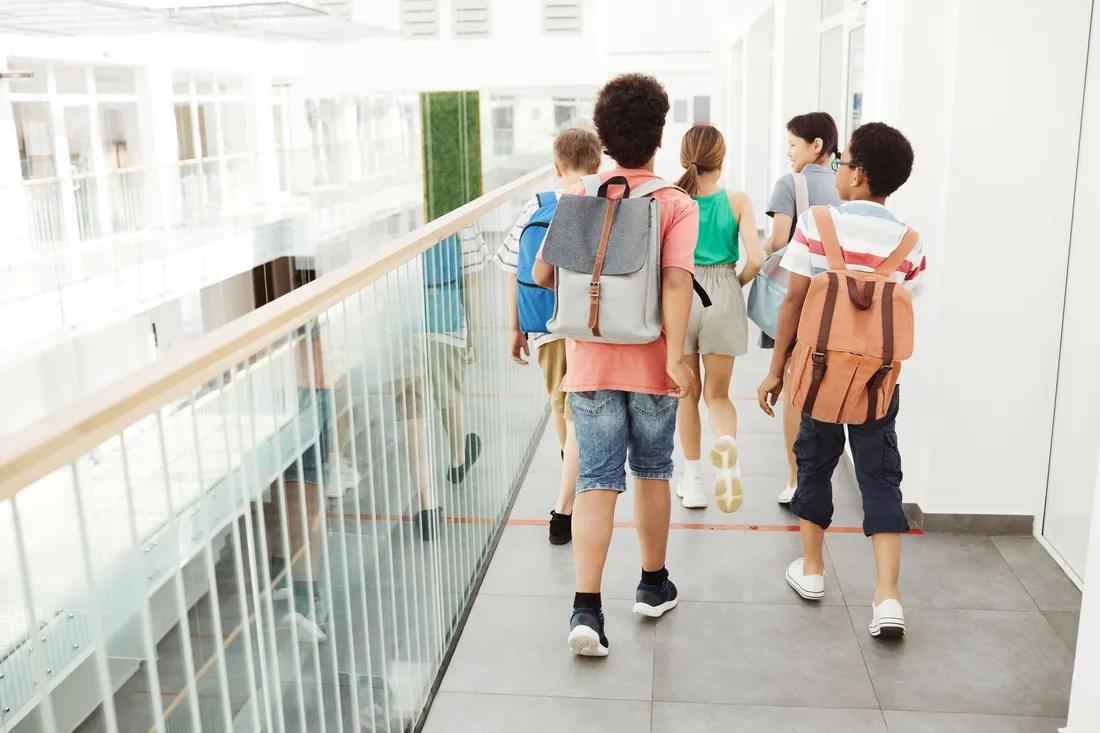 Des collégiens portant des sacs à dos marchent de dos dans la coursive vitrée d'un établissement scolaire neuf, sol carrelé gris et garde-corps en verre