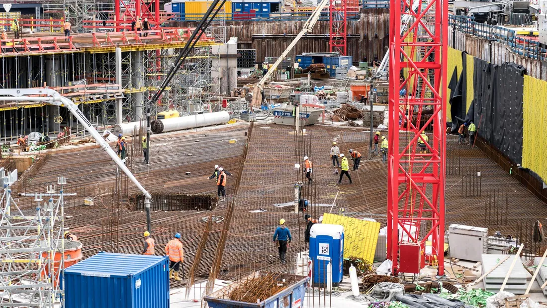 Chantier de bâtiment au sol : dalle en cours de ferraillage, pompe à béton, grue à tour rouge et ouvriers en gilets de sécurité au travail