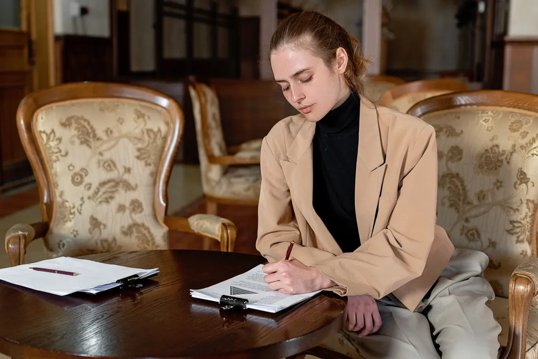 Une femme en veste beige, assise à une table ronde en bois dans une pièce meublée de fauteuils anciens, lit et annote au stylo un dossier de plusieurs pages tenu par une pince noire ; un second jeu de feuilles et un stylo sont posés à côté sur la table