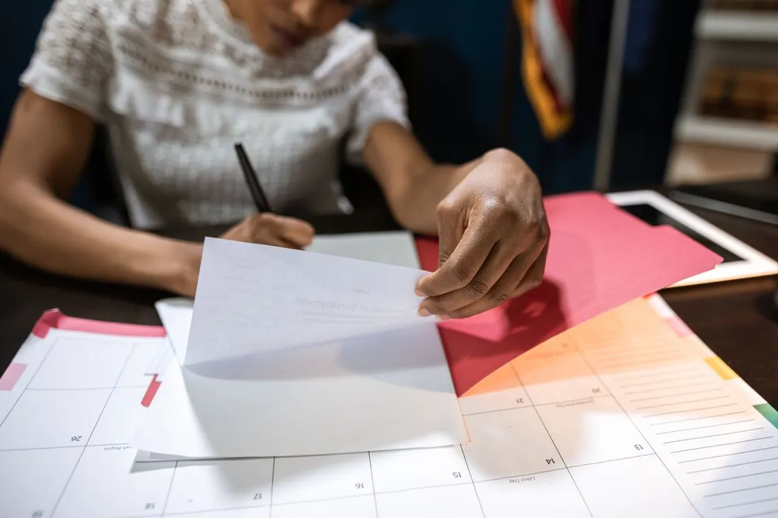 Mains d'une femme posant une feuille de papier sur un grand calendrier mensuel de bureau, à côté de chemises cartonnées de couleur et d'un stylo