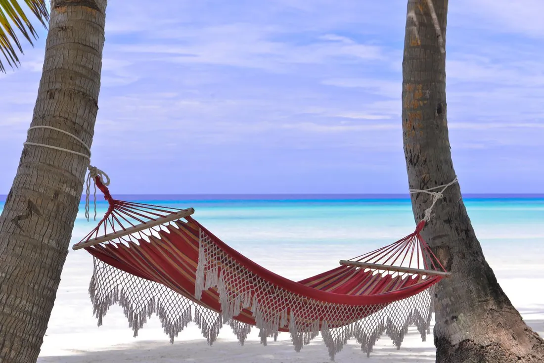 Hamac rouge tendu entre deux cocotiers sur une plage de sable blanc face a une mer turquoise, evoquant le repos pour mieux recuperer du decalage horaire