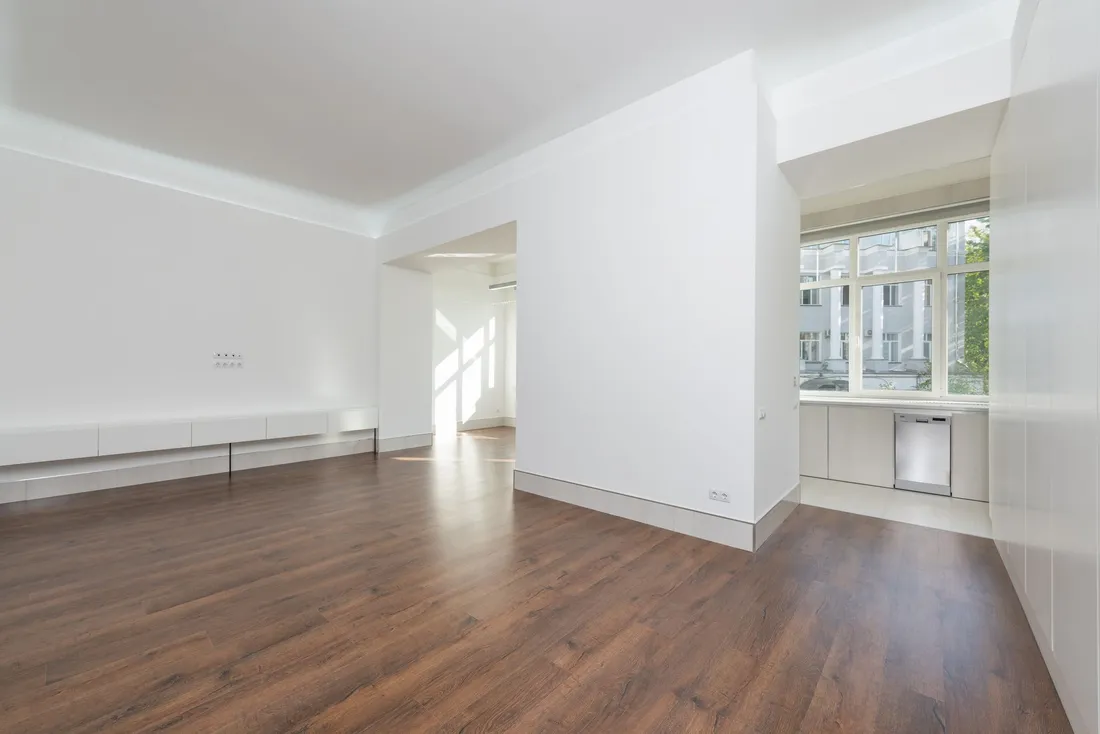Séjour d'appartement entièrement vide, murs blancs, parquet sombre et grande fenêtre donnant sur un immeuble voisin, sans aucun meuble