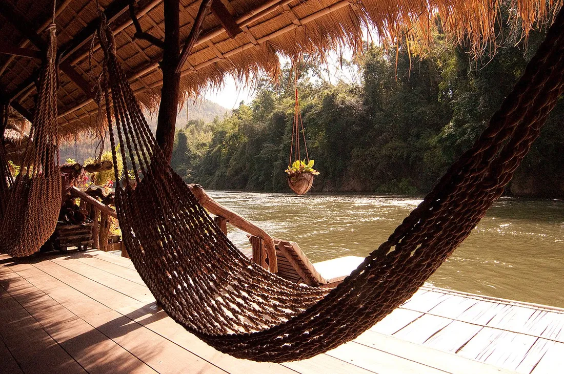 Hamacs tresses suspendus sous un carbet au toit de palmes, face a une riviere bordee de foret tropicale