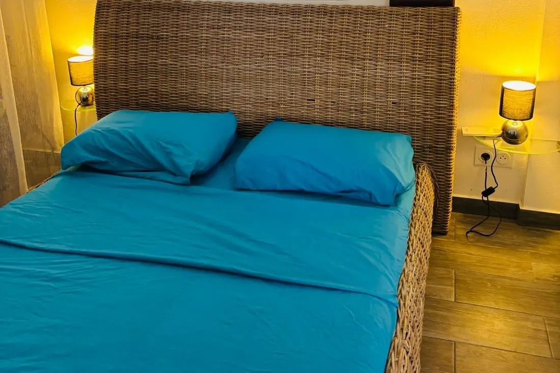Chambre de la maison Tropikana : lit double à tête en rotin, parure turquoise et lampes de chevet allumées