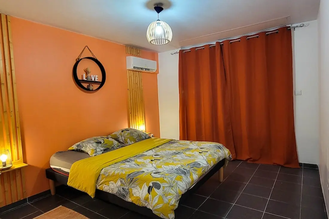 Chambre de Cayenne Waouh : mur orange, grand lit à housse jaune à motifs de toucans, rideau orange et climatisation