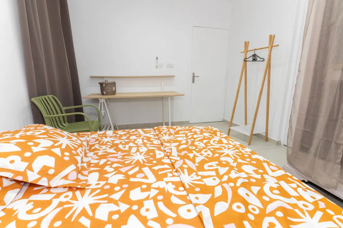 Chambre du Nid de Thalys : lit à housse orange à motifs, bureau en bois clair avec chaise verte et portant en bambou