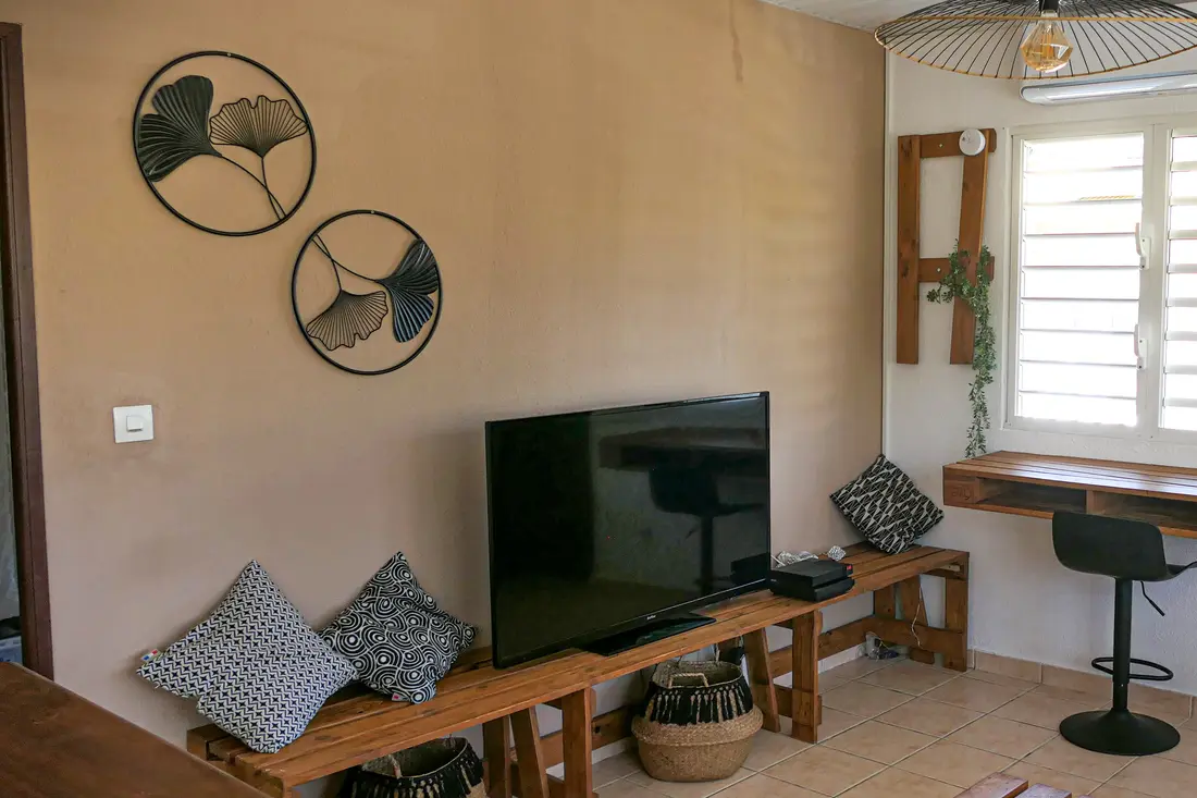 Séjour d’un appartement Cayenne Chic : banc en palettes avec coussins graphiques, télévision et bureau suspendu près de la fenêtre