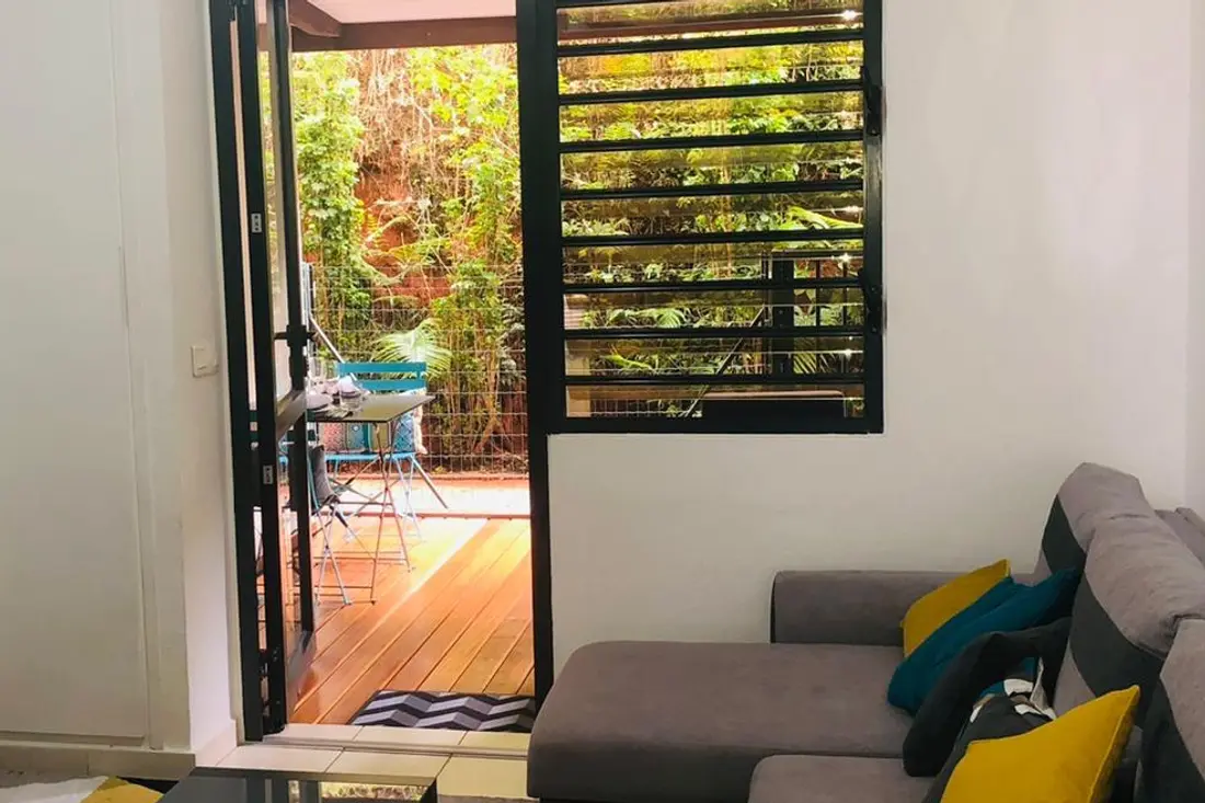 Séjour du Diamant Cayennais ouvert sur sa terrasse en bois : canapé d’angle à coussins jaunes et table basse, verdure dense derrière les jalousies