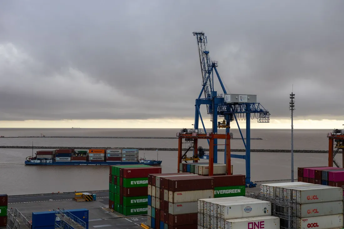 Quai d'un port de commerce vu du sol : portique à conteneurs bleu et rouge, piles de conteneurs sur le terre-plein et caboteur chargé de conteneurs sur un fleuve aux eaux brunes