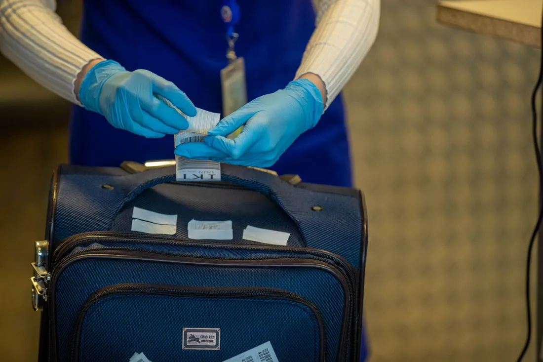 Mains gantées de bleu d'un agent en gilet bleu marine manipulant l'étiquette à code-barres d'une valise posée sur un comptoir de contrôle