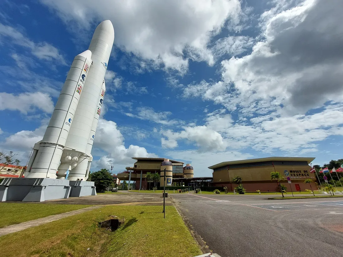 Entrée du Centre spatial guyanais à Kourou : maquette grandeur nature d'une fusée Ariane 5 dressée sur son socle, bâtiment d'accueil vitré en arrière-plan et halle du Musée de l'espace sur la droite, devant un parking désert