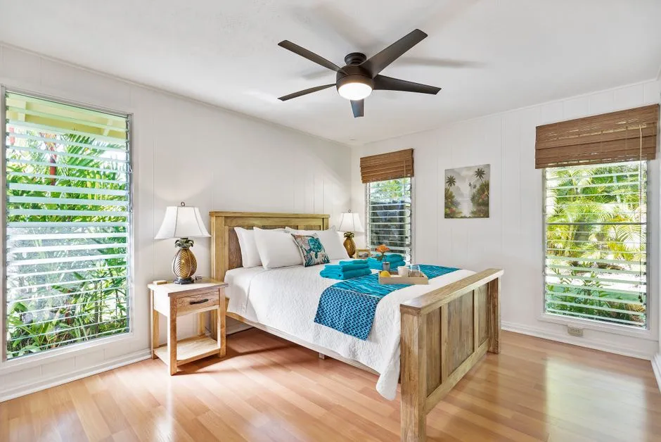 Chambre lumineuse d'une location de vacances tropicale avec ventilateur de plafond, jalousies et persiennes en bois ouvertes sur la vegetation, mobilier en bois clair