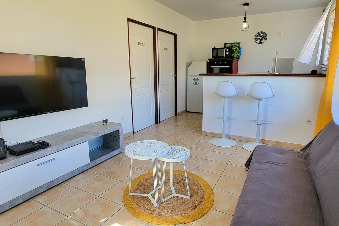 Séjour d’un appartement Cayenne Chic : canapé gris, tables basses blanches, coin cuisine avec bar et tabourets au fond