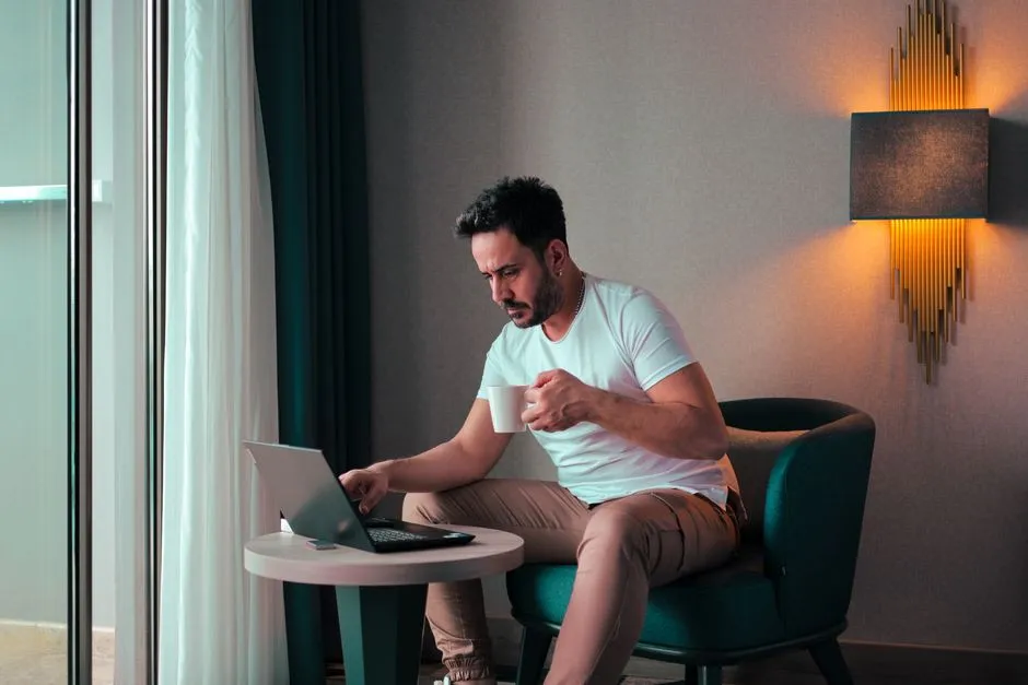 Voyageur d'affaires installé dans un fauteuil de sa chambre d'hébergement, une tasse de café à la main, consultant son ordinateur portable posé sur une table basse