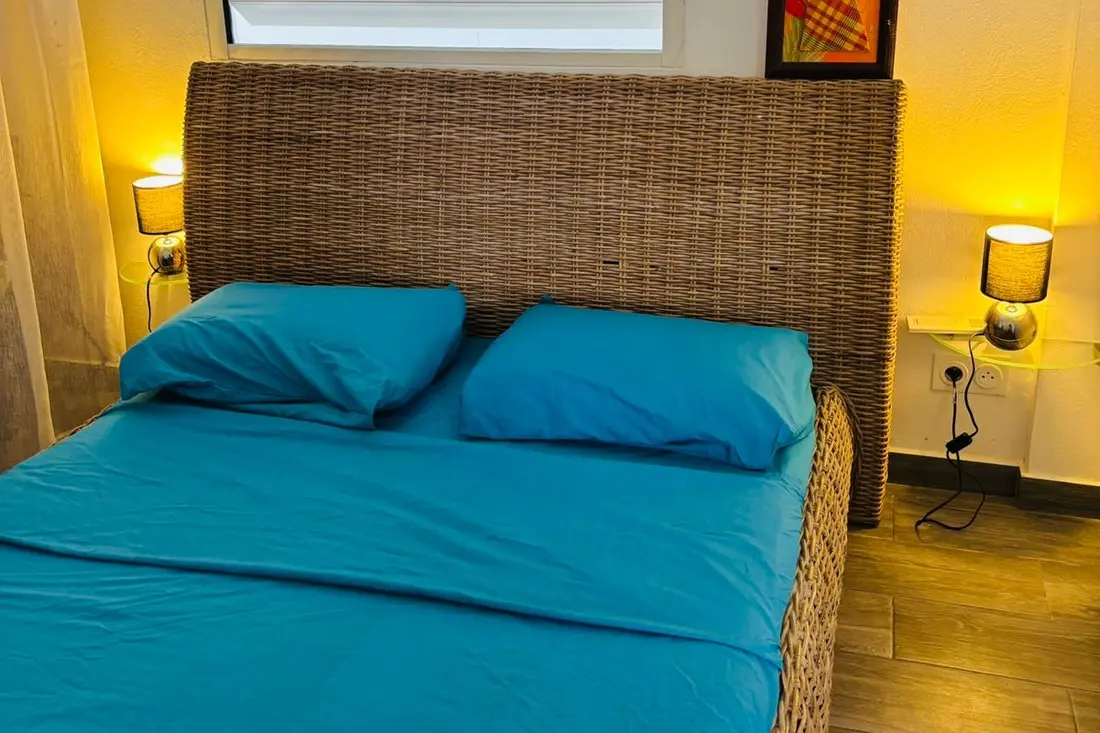 Chambre de la Tropikana à Rémire-Montjoly : lit en rotin à parure bleu canard entre deux appliques allumées