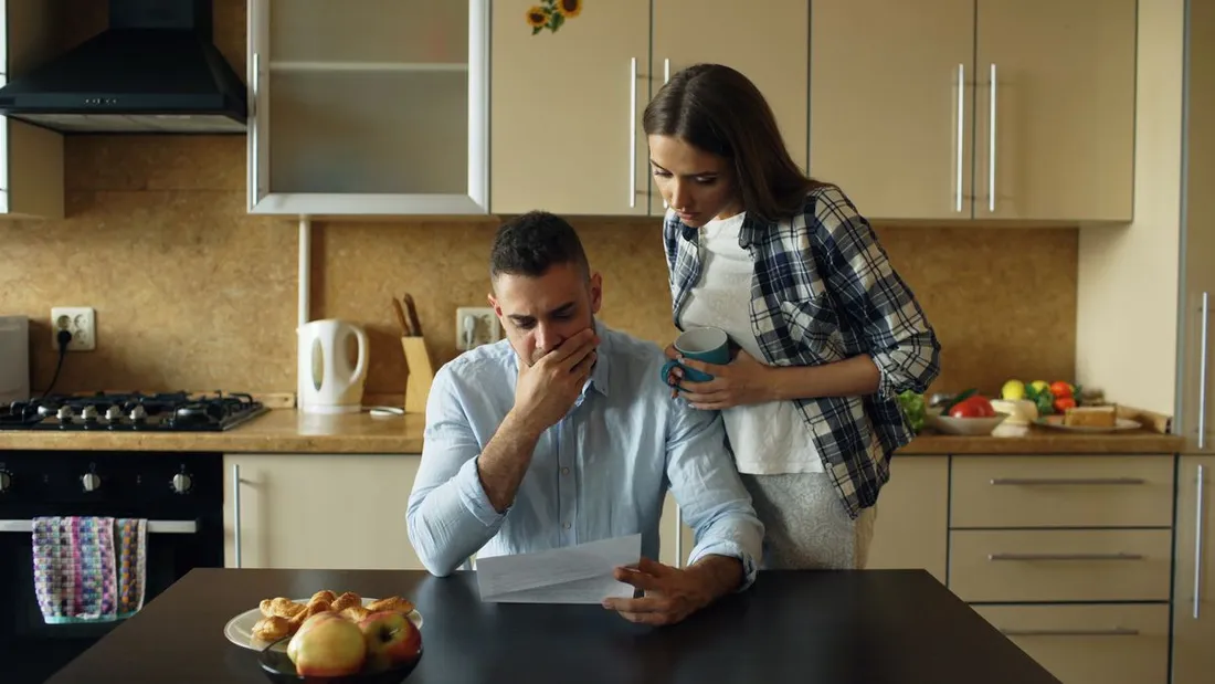 Dans une cuisine, un homme assis à table lit une lettre d'un air soucieux, la main devant la bouche, une femme debout derrière lui lit par-dessus son épaule en tenant une tasse