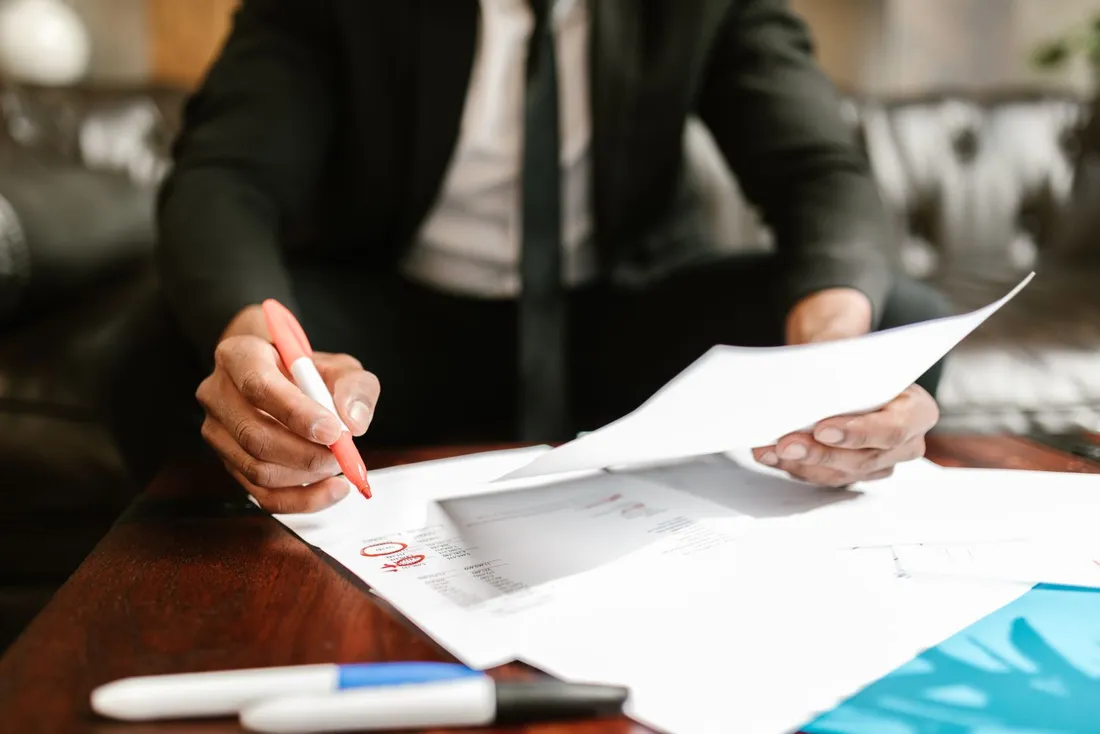 Une personne en veste sombre, assise à une table en bois, relit un document imprimé qu'elle tient d'une main et annote une autre feuille au stylo rouge