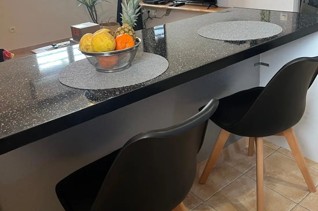 Bar de cuisine d’un appartement Cayenne Chic : plan noir pailleté, corbeille de fruits et deux chaises scandinaves noires