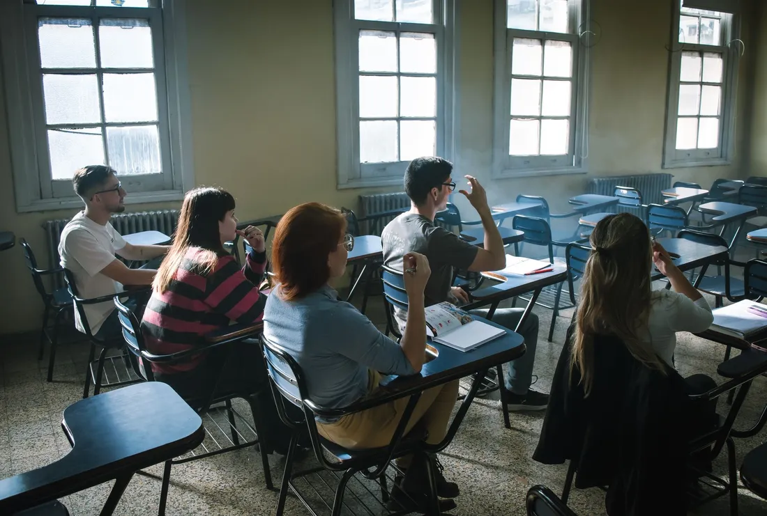 Adultes assis à des tables-chaises dans une salle de cours claire, cahiers et manuels ouverts, l'un d'eux levant la main vers le formateur hors champ