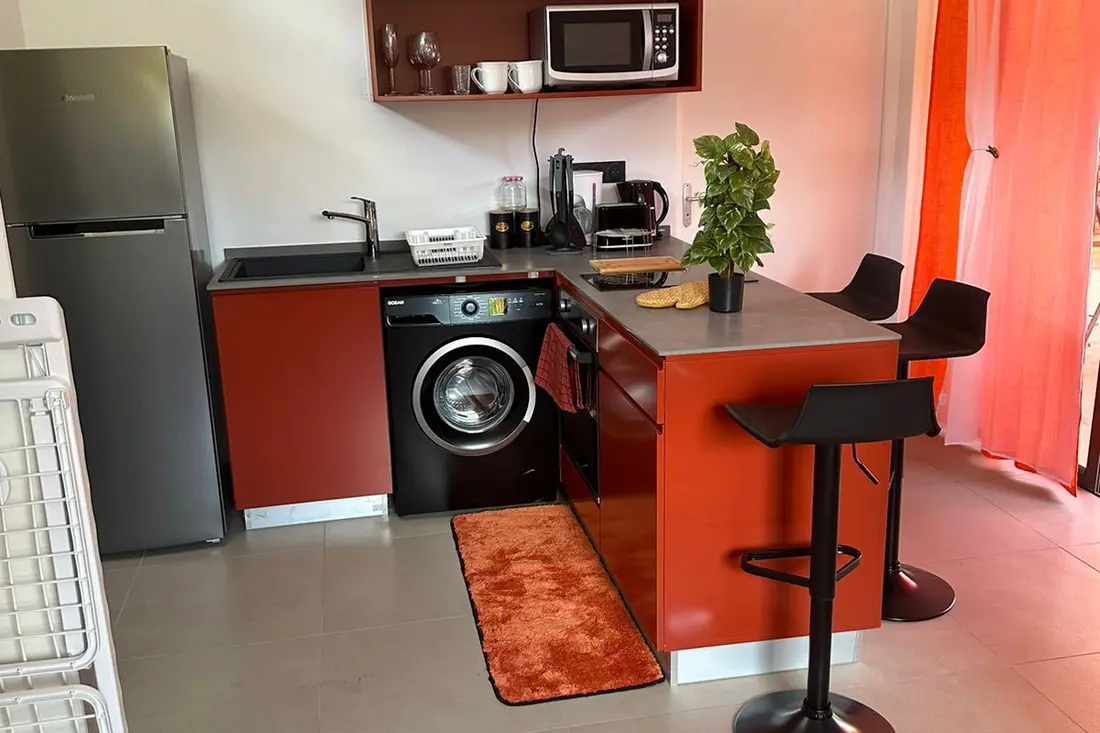 Cuisine rouge d’un studio de la Résidence Francis : lave-linge noir, bar avec tabourets et plante sur le comptoir gris