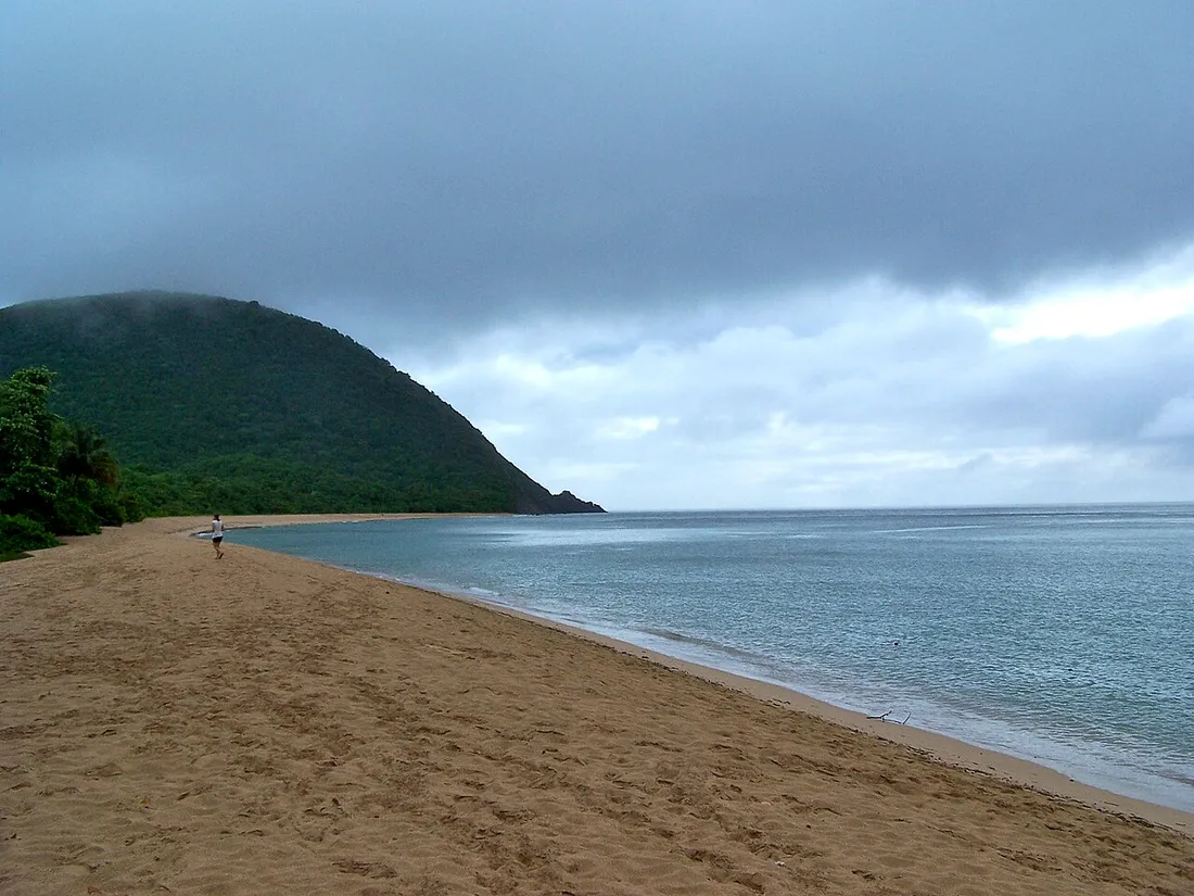 Une personne seule marchant sur la plage de Grande Anse en Guadeloupe, mer calme et collines verdoyantes en arrière-plan