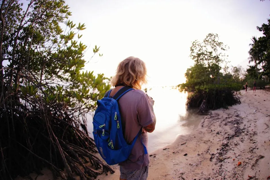 Voyageur solo avec sac à dos bleu sur une plage tropicale bordée de mangrove et de palmiers au coucher du soleil