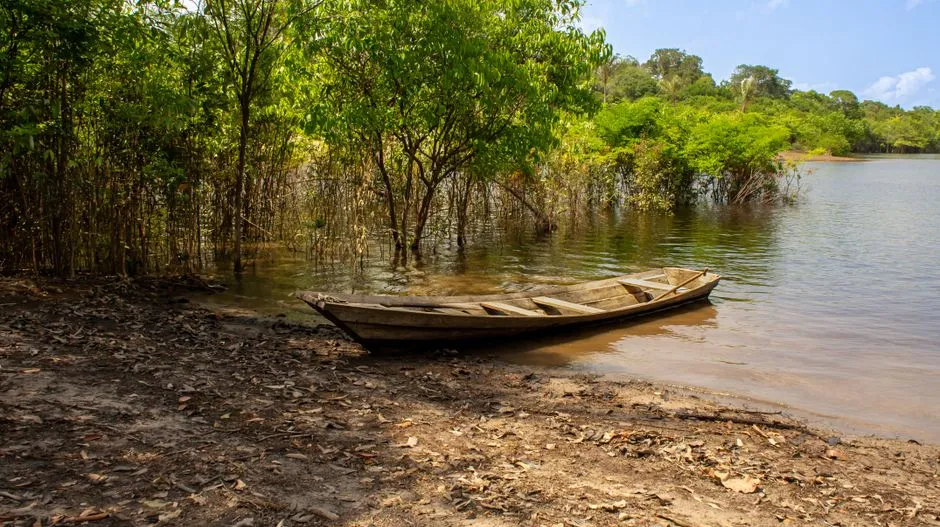 Pirogue en bois amarree sur la berge d'un fleuve borde de foret amazonienne, evoquant les excursions et les distances a parcourir en Guyane