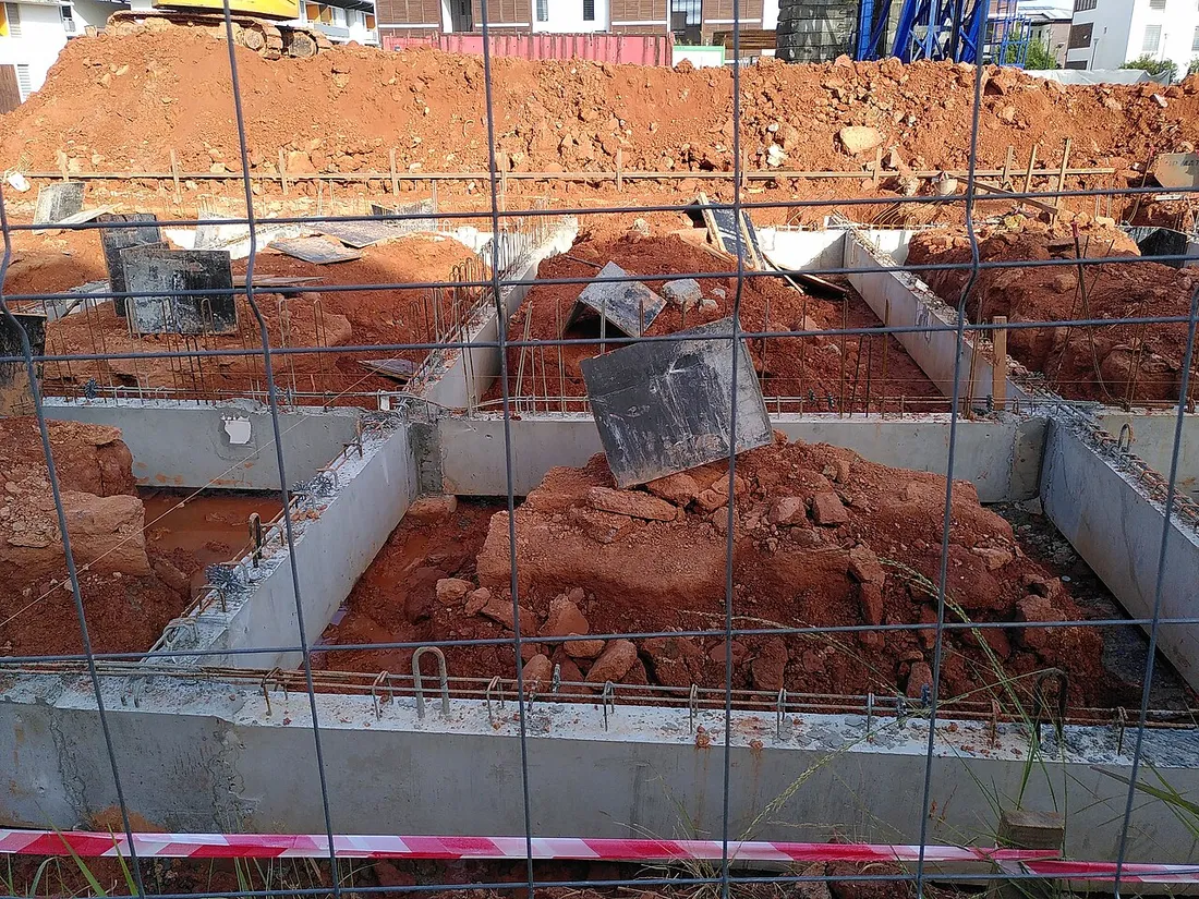 Chantier de construction à Cayenne en Guyane : fondations en béton et terre latéritique rouge derrière une clôture de sécurité