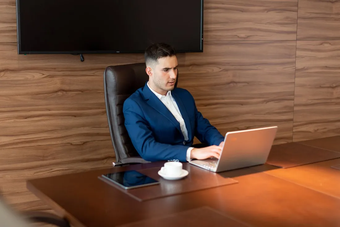 Consultant en costume bleu concentré sur son ordinateur portable dans une salle de réunion moderne, tasse de café posée sur le bureau