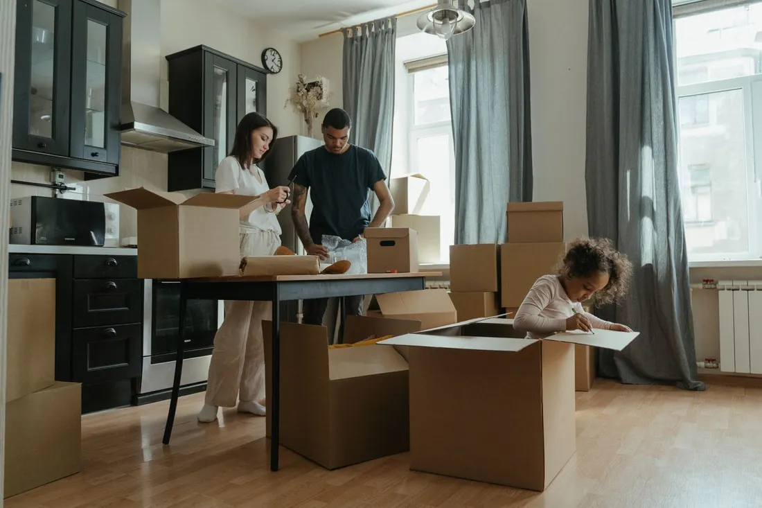 Un couple et leur jeune enfant déballent des cartons de déménagement dans la cuisine d'un appartement, la petite fille dessinant sur un carton
