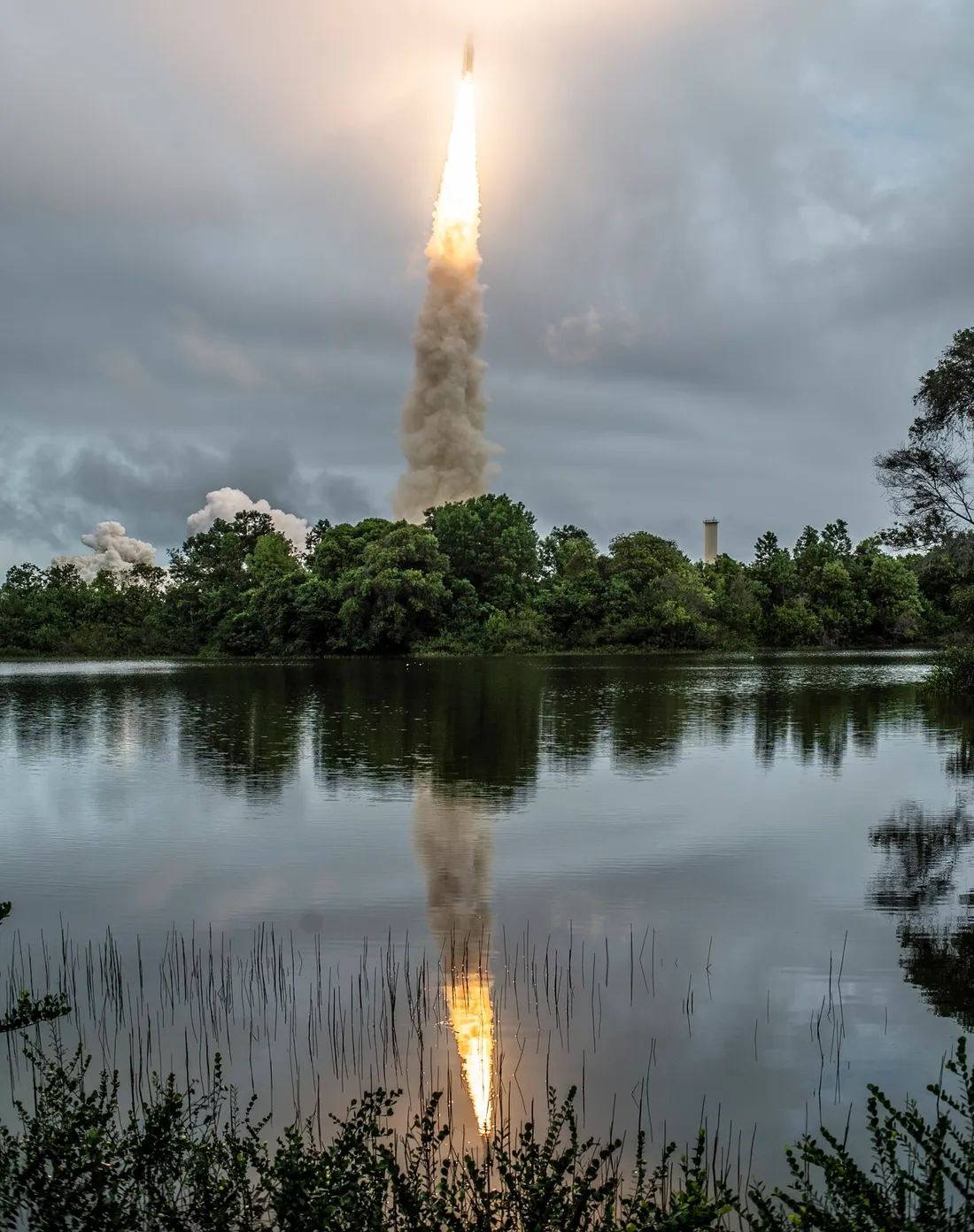 Décollage d'une fusée Ariane 5 depuis la zone de lancement ELA-3 du Centre spatial guyanais à Kourou, la colonne de flammes et de fumée se reflétant dans un plan d'eau bordé de forêt