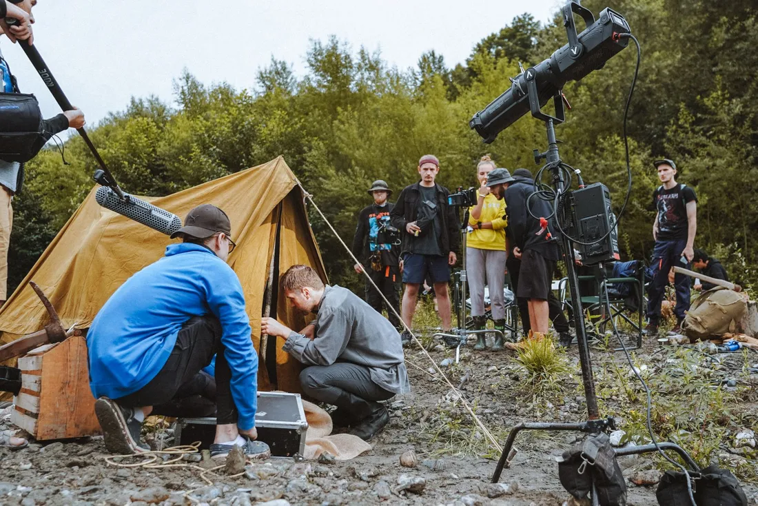Équipe de tournage installée en extérieur au bord d'une rivière, avec projecteur sur pied, perche son et caméra, pendant une journée de prises de vues en pleine nature