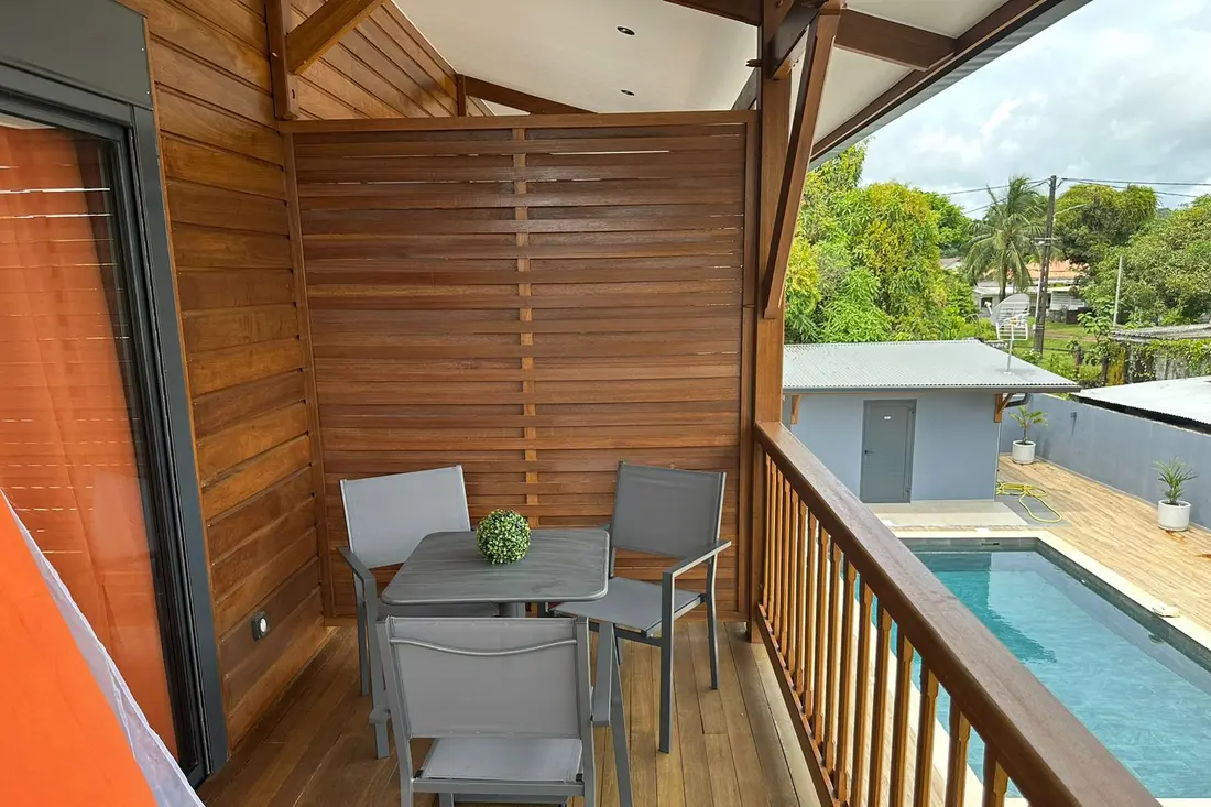 Balcon en bois de la Résidence Francis à Cayenne : table et chaises de jardin, piscine de la résidence en contrebas
